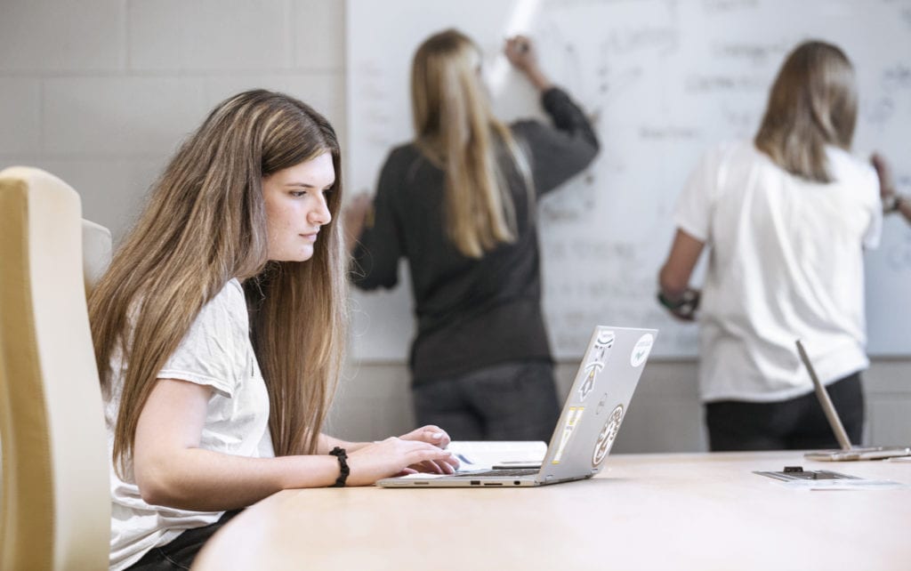 two women writing on white board while another woman is on a laptop