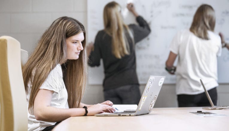 two women writing on white board while another woman is on a laptop